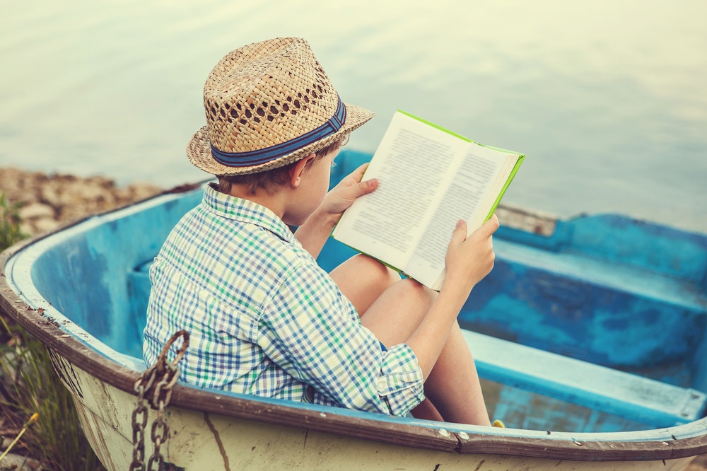 Kafasında şapka olan bir çocuk küçük bir teknede kitap okuyor.
