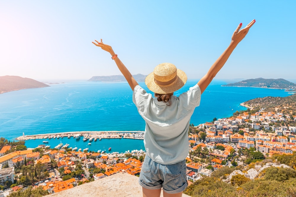 A happy woman with open arms stands on a viewpoint enjoying the panorama of Kas resort on the Mediterranean Sea in Turkey.