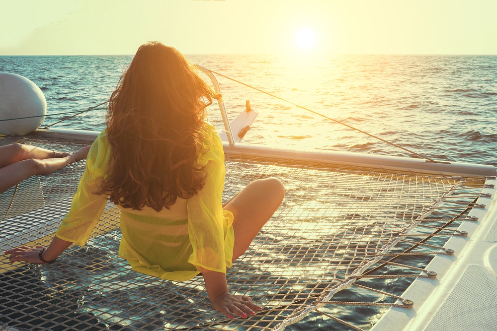 A woman sits on a net on a catamaran, watching the sunset with one.
