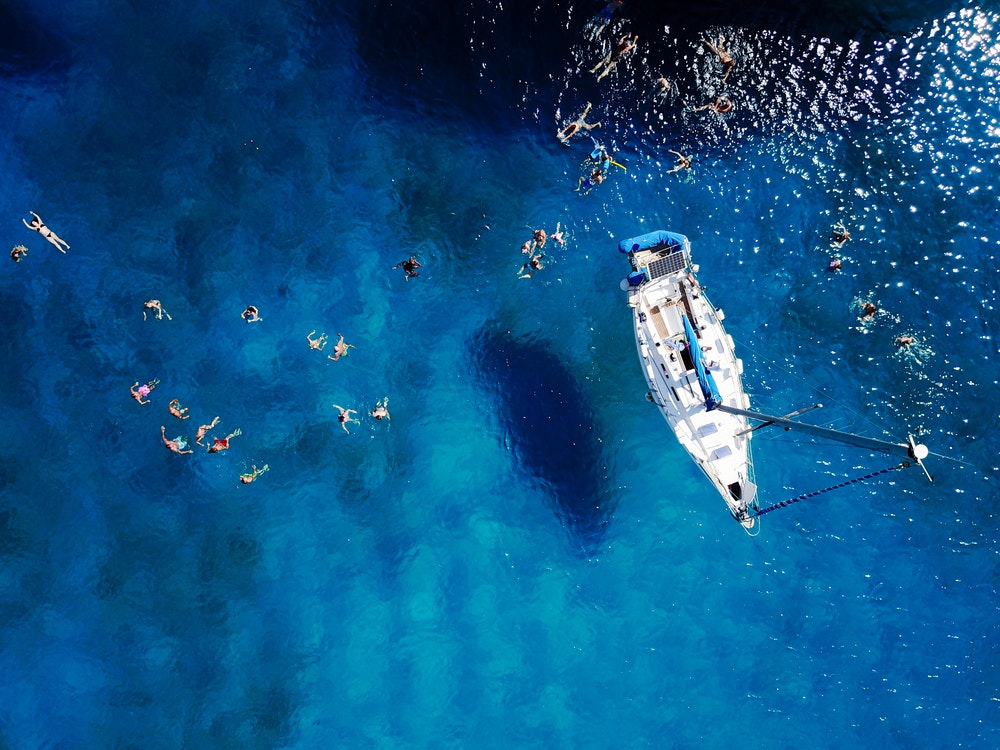 Karštą vasaros dieną iš oro nufotografuota graži mėlyna lagūna su burlaiviu. Žmonių, plaukiojančių aplink valtį, vaizdas iš viršaus.