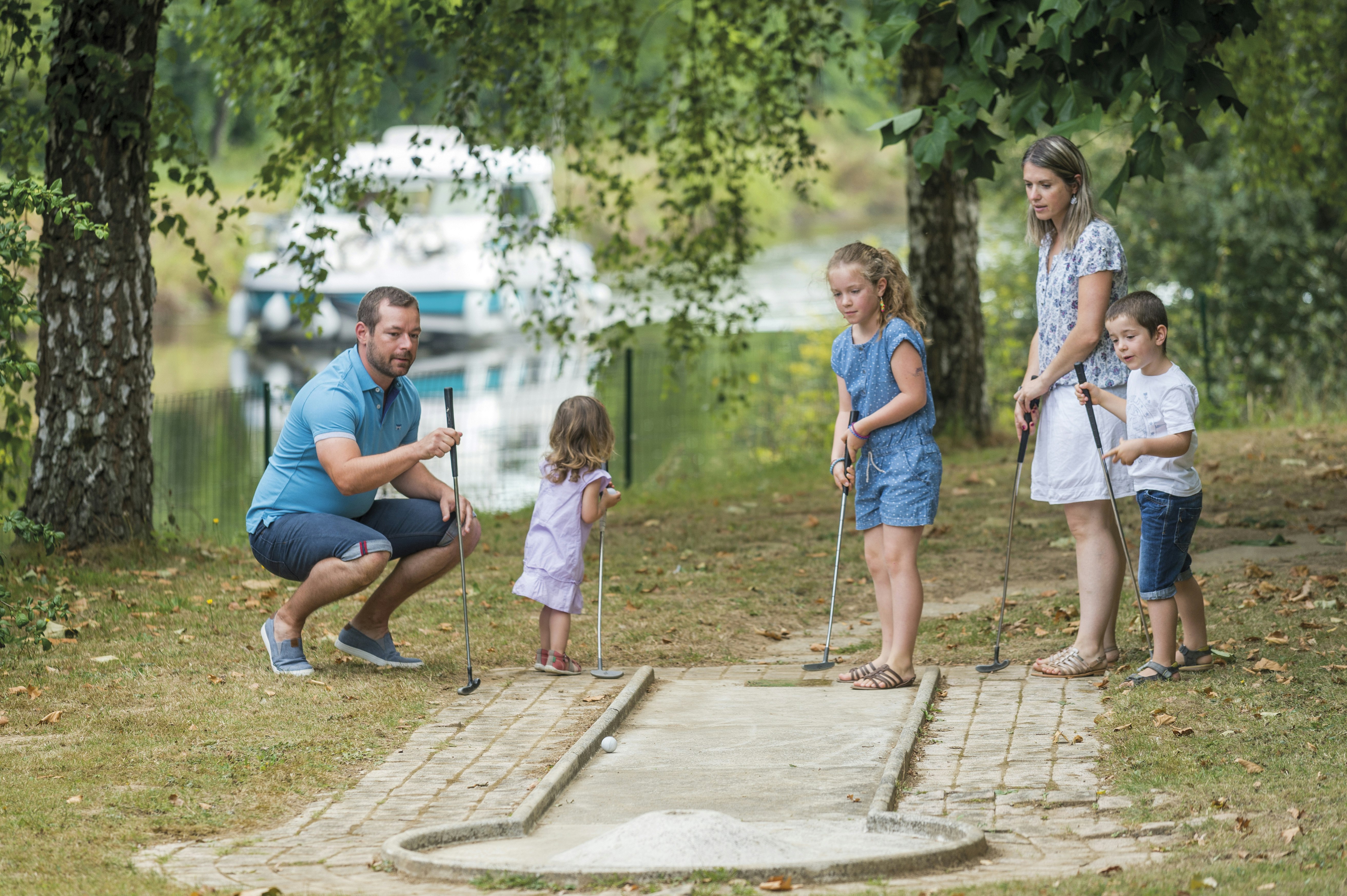 Rodina hrající minigolf s vodním kanálem a hausbótem v pozadí