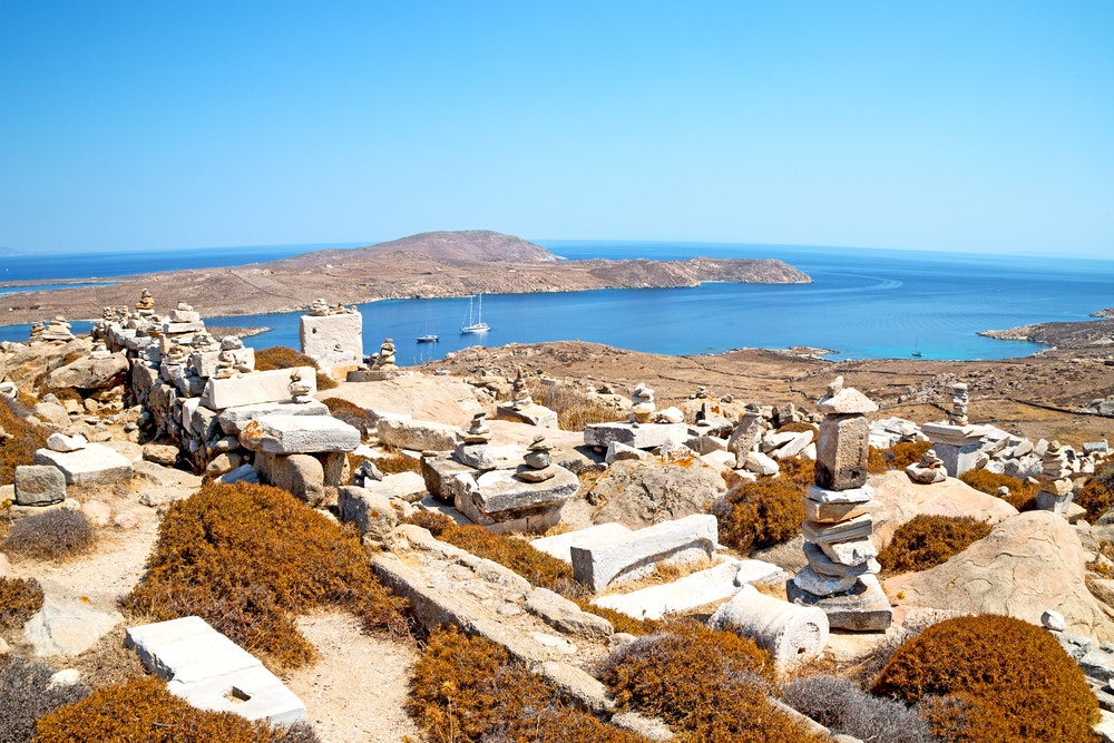 Archaeological monument on the island of Delos in the background with a bay and a yacht.