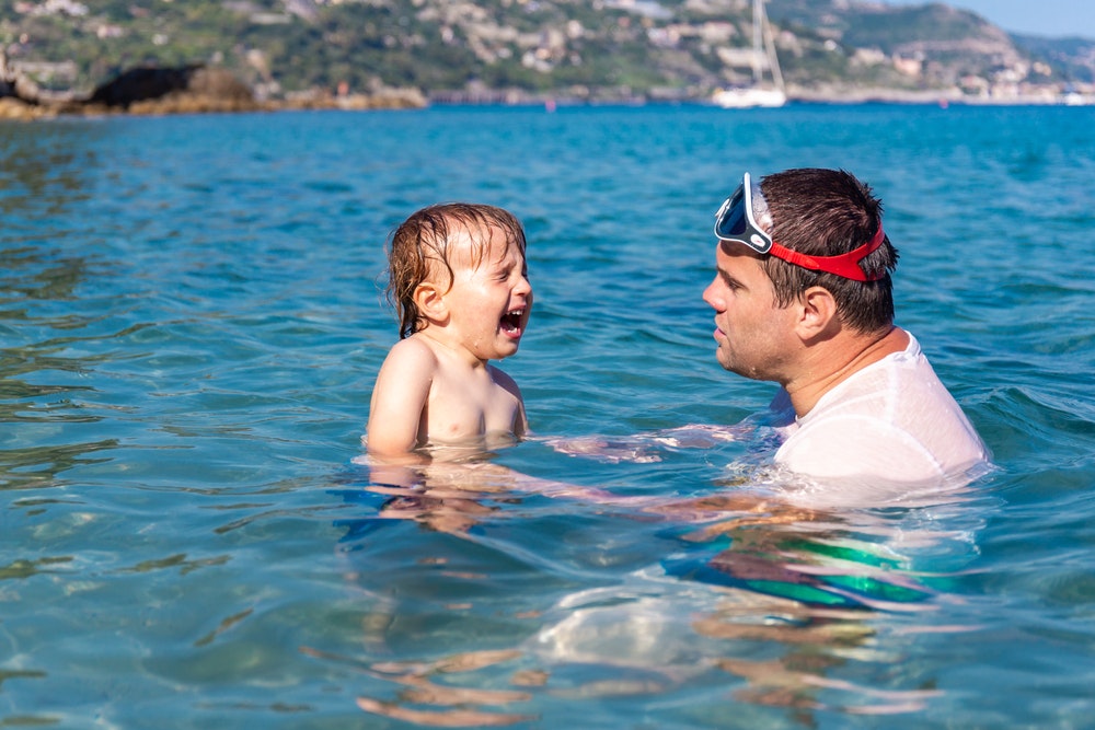 A small child cries and is afraid of the water in the sea. Father and son swimming together in the sea during the summer holidays.