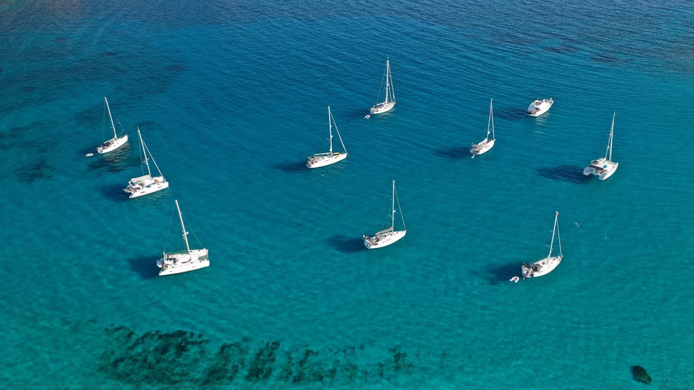 Blick von oben auf eine türkisfarbene Meeresbucht mit weit auseinander liegenden, vor Anker liegenden Segelbooten.
