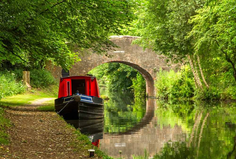 Ein Hausboot auf dem ruhigen Wasser eines Kanals in England mit einer Steinbogenbrücke im Hintergrund.