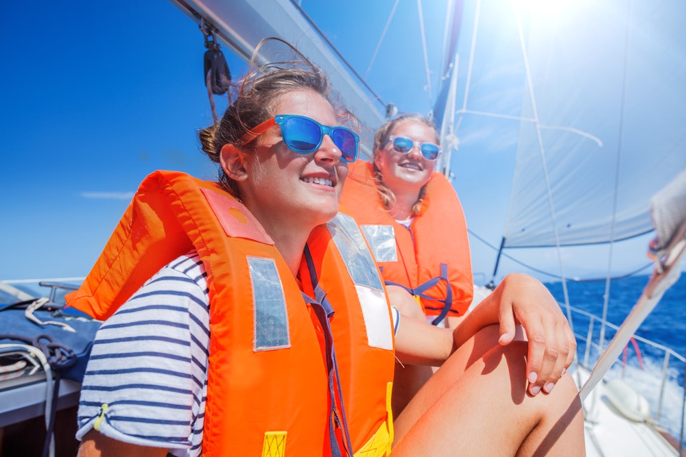 Girls on board a sailboat wearing life jackets and sunglasses.