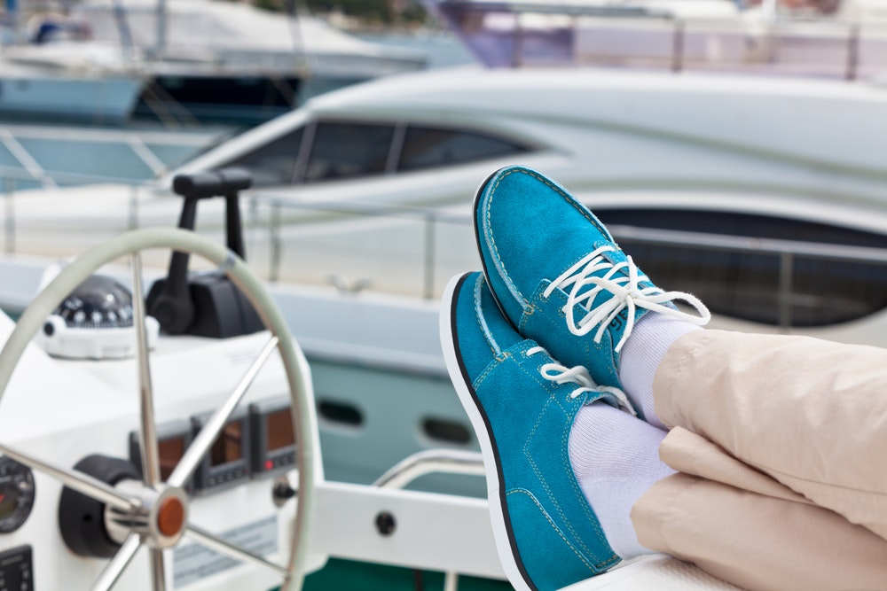 A pair of human legs in trousers and bright blue boat shoes against the backdrop of a yacht